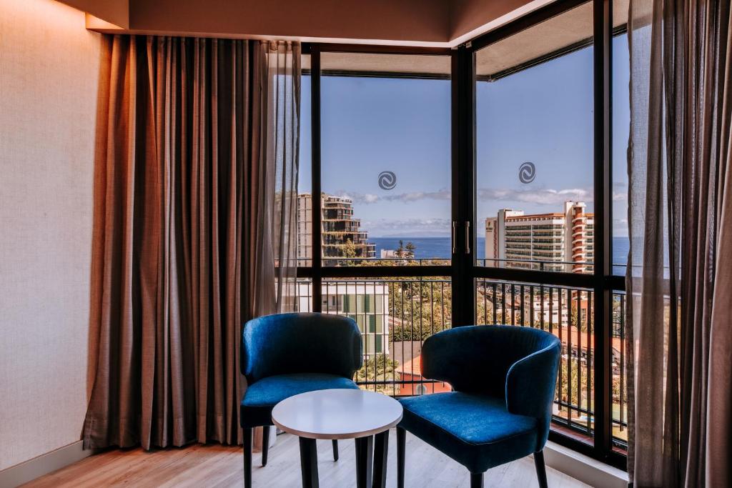 Mūsdienīgs interjers ar zilu krēslu un baltu galdiņu, kas izvietots pie liela loga ar skatu uz pilsētu un jūru. Dabiska gaisma piepilda telpu, radot patīkamu atmosfēru.