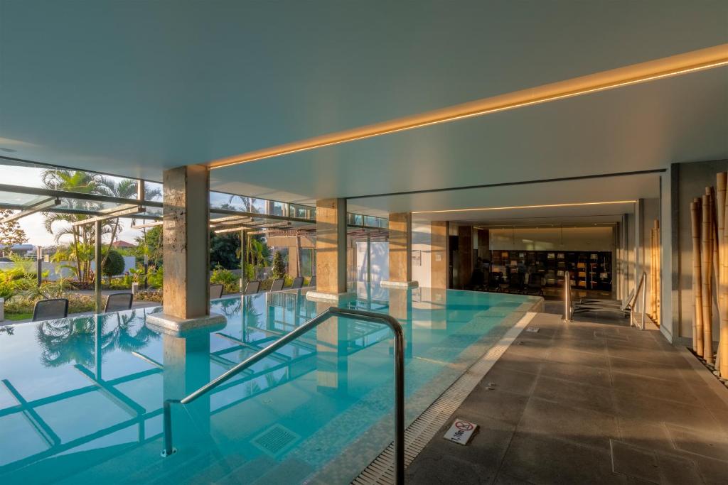 Mūsdienīgs iekštelpu baseins ar skatu uz dārzu, apgaismots ar siltām gaismām un ar marmora akcentiem. Apkārt ir augi un ērtas sēdvietas, radot relaksējošu atmosfēru.