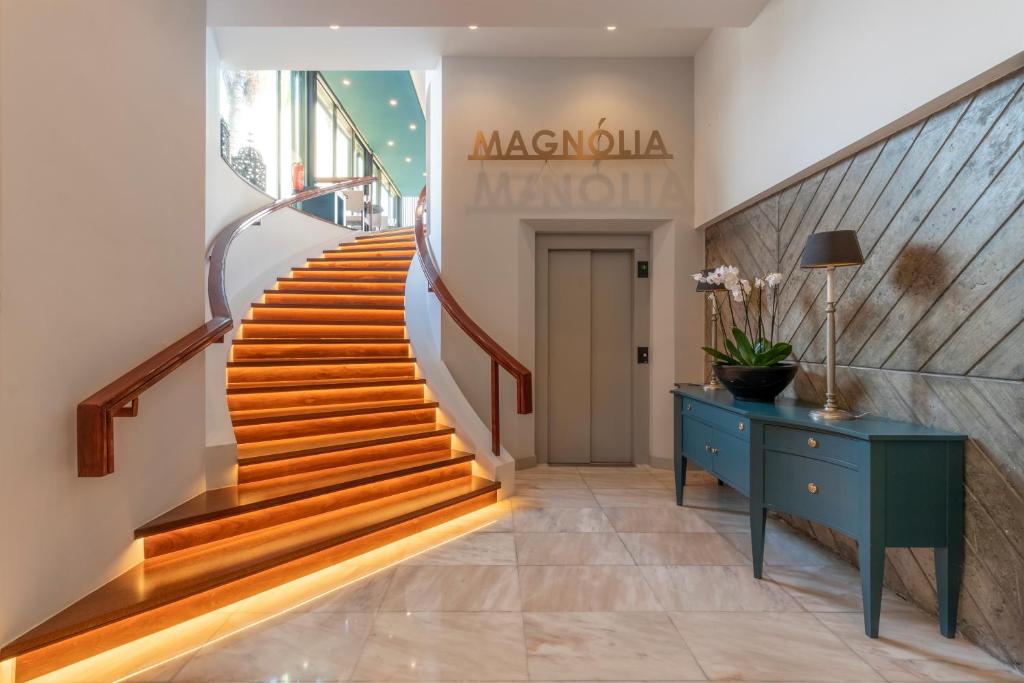 Eleganta kāpnes ar apgaismojumu, kas ved uz augšu, ar modernu interjeru un viesmīlīgu uzņemšanas zonu. Telpā redzams stilīgs galds ar ziediem un liftu.