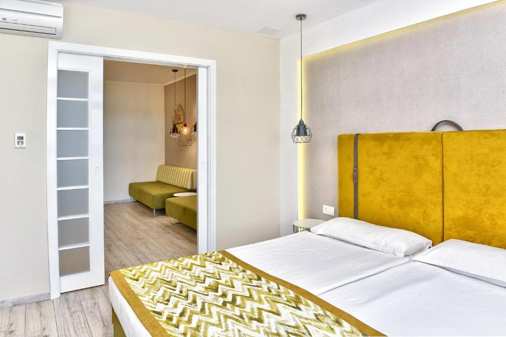 Mūsdienīga guļamistaba ar dzeltenu gultas galvu un stilīgu dekoru. Caur stikla durvīm redzama atpūtas zona ar zaļiem dīvāniem.
