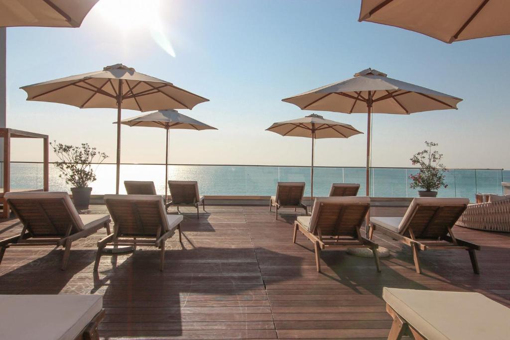 Sauļošanās krēsli ar saulessargiem pie jūras, radot relaksējošu atmosfēru. Skaists skats uz ūdeni un sauli, kas noriet.