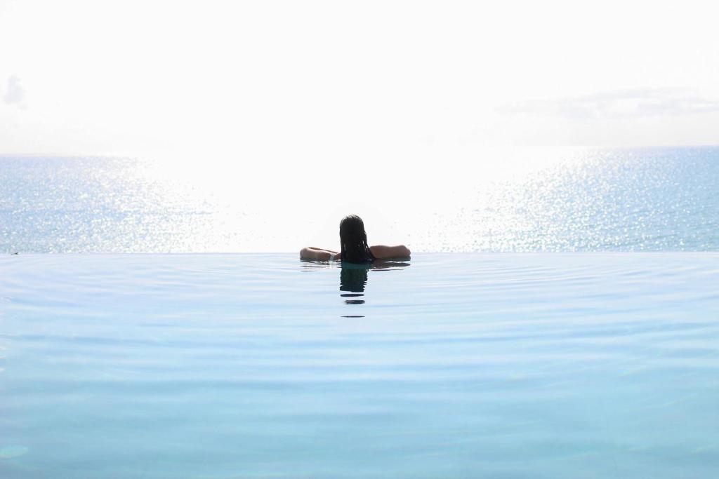 Mierīgi cilvēks atpūšas bezgalīgā baseinā, skatoties uz saulrietu, kas atspoguļojas ūdenī.