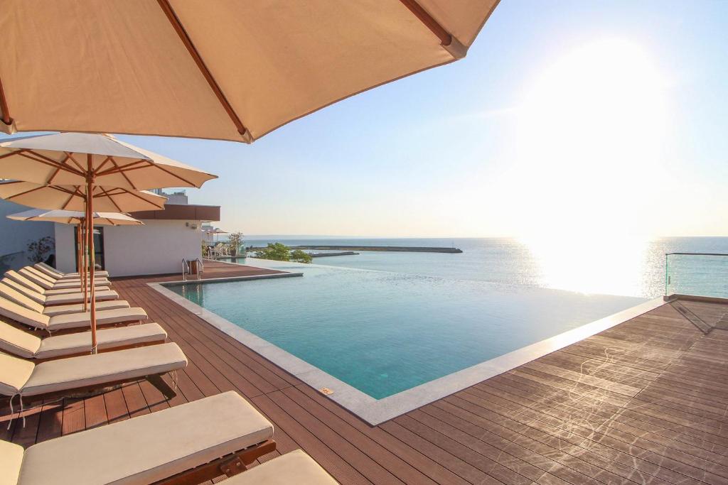 Luksusīgs baseina skats ar sauļošanās krēsliem un saulessargiem, kas atrodas pie jūras. Aiz baseina paveras gleznains horizonts un spilgta saule.