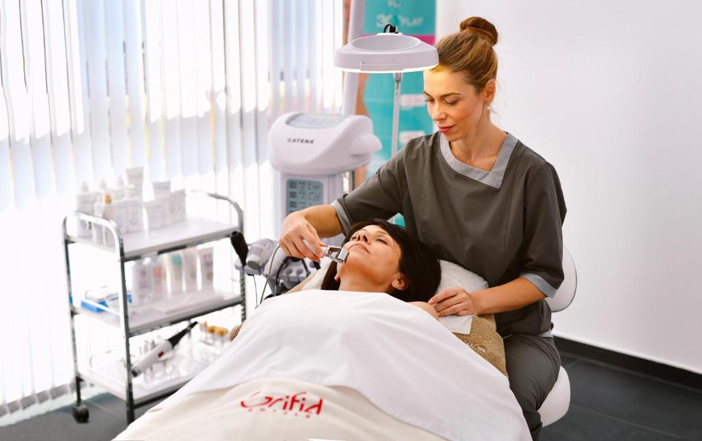 Skaistumkopšanas procedūra, kurā kosmetologs veic sejas ārstēšanu ar modernu aprīkojumu. Kliente guļ uz gultas, atsvaidzinoša atmosfēra.