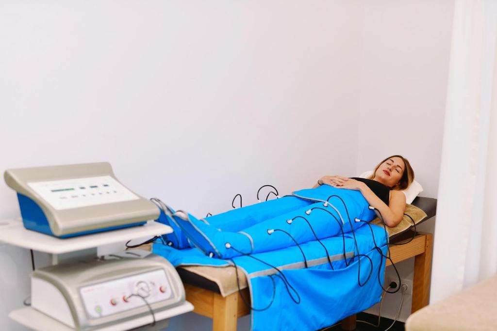 Sieviete guļ uz ārstniecības gultas, apsegtā ar zilu segu un pieslēgta pie medicīnas iekārtām, kas tiek izmantotas terapijā. Telpa ir gaiša un vienkārša, radot relaksējošu atmosfēru.