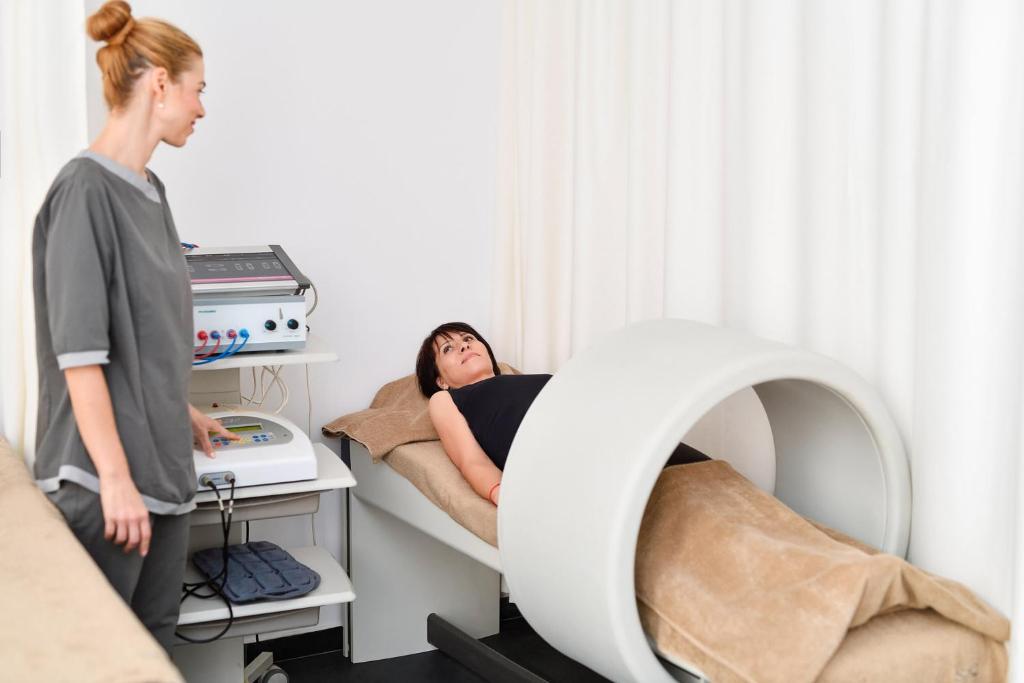 Apspriežot fizioterapiju, sieviete guļ uz terapijas galda ar ierīci blakus, kamēr speciāliste veic procedūru.