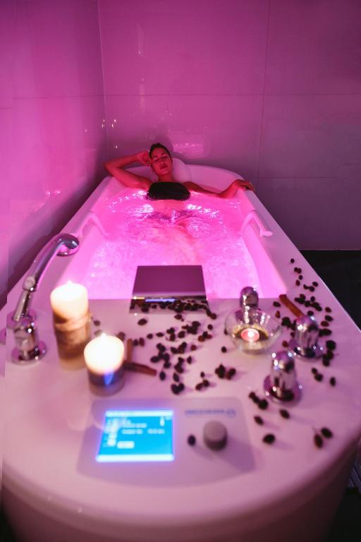 Relaksējoša vanna ar rozā gaismu, kurā atpūšas sieviete, apkārt sveces un rožu ziedlapiņas. Ideāla vide mierīgai un romantiskai atmosfērai.