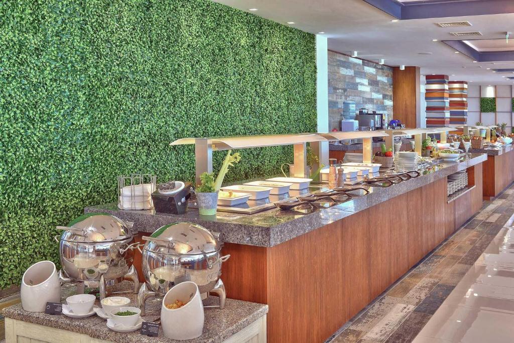 Mūsdienīga restorāna bufete ar zaļo augu sienu, kurā ir dažādi ēdieni un dzērieni. Eleganti pasniegti trauki un gaļas sautēšanas katli.