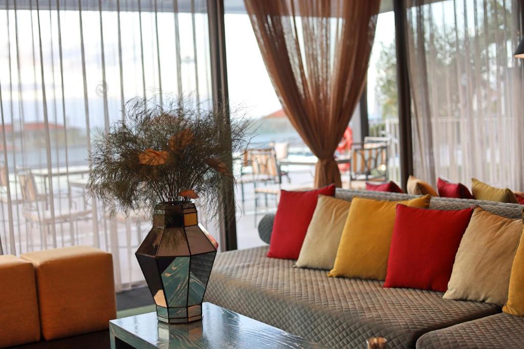 Mūsdienīgs dzīvojamās telpas interjers ar krāsainām spilveniem, elegantu ziedu podu un caurspīdīgām aizkariem, kas rada mājīgu atmosfēru.