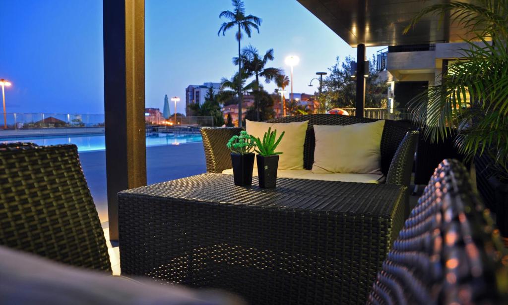 Mūsdienīgs āra atpūtas stūris ar ratanveida mēbelēm, galda centrā atrodas puķu pods ar kaktusu. Fons ietver baseinu un pilsētas apgaismojumu vakara zilmē.