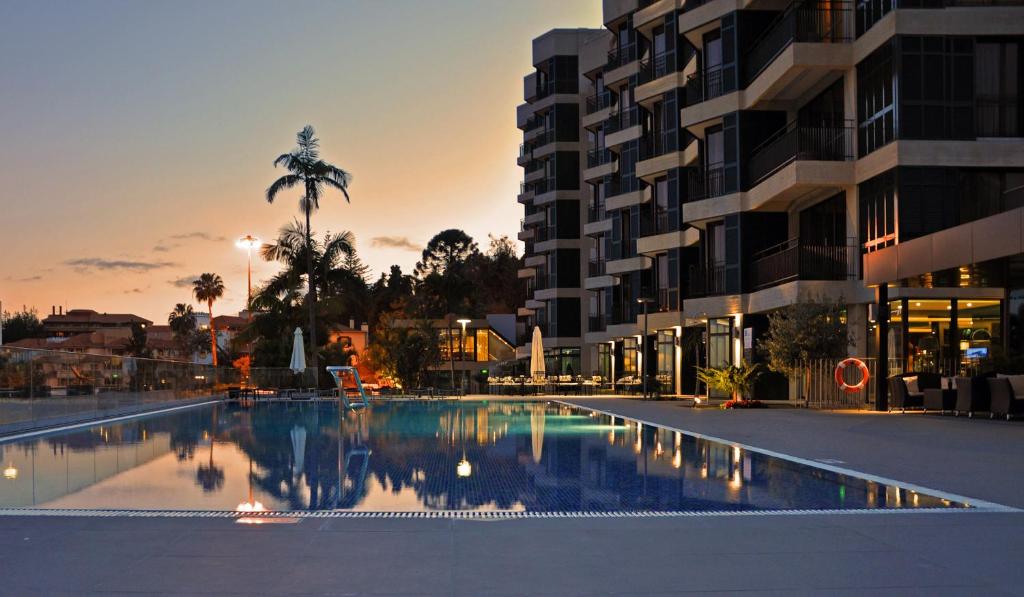 Eleganta viesnīca pie peldbaseina saulrieta gaismā, ar palmām un mūsdienīgu arhitektūru. Ūdens virsma atspoguļo apkārtējo ainavu, radot mierīgu atmosfēru.