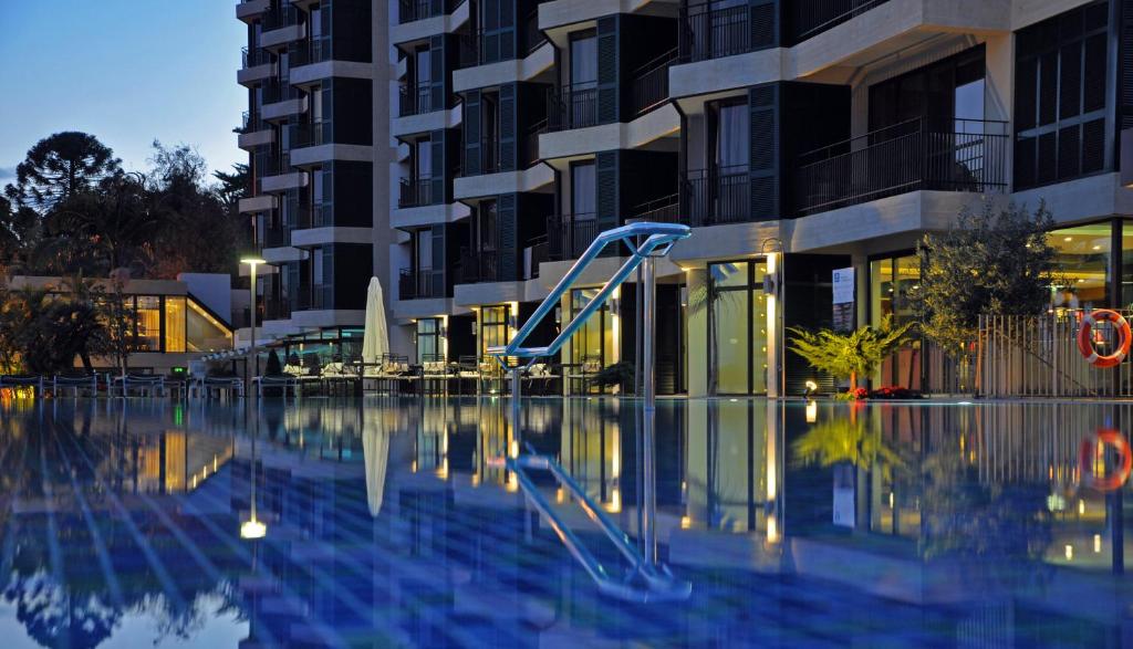 Moderns dzīvokļu komplekss ar baseinu, kas atspoguļojas ūdenī, un slidkalniņu, kas atrodas pie ēkas. Apgaismojums radījis patīkamu atmosfēru vakara stundās.