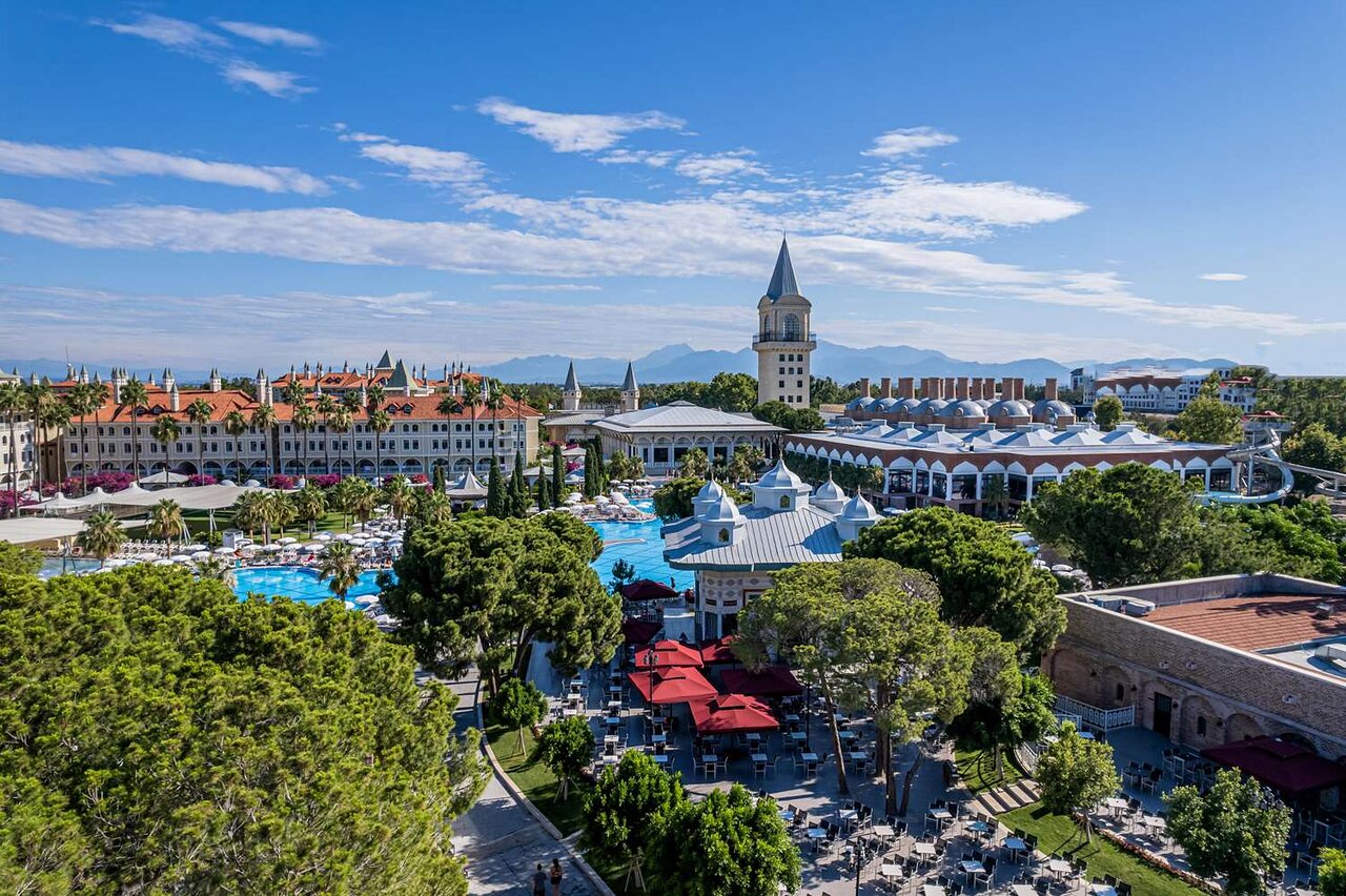 Liels viesnīcas komplekss ar peldbaseinu un skaistiem dārziem zem saules, kas spīd uz debesīm. Apkārt ir zaļas koka un moderni arhitektūras elementi. panoramic view with beach and pools