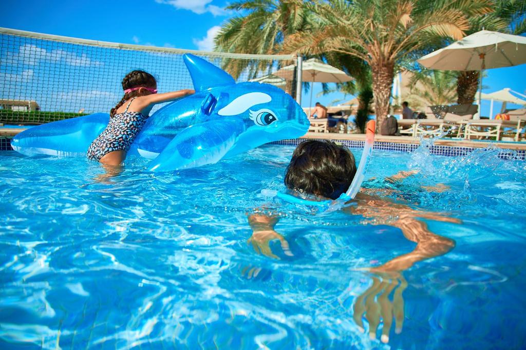 Bērni spēlējas baseinā ar zilu delfīna peldriņķi, kamēr viens no viņiem snorkelē. Apkārt ir saulaina atmosfēra ar palmām un atpūtas vietām.