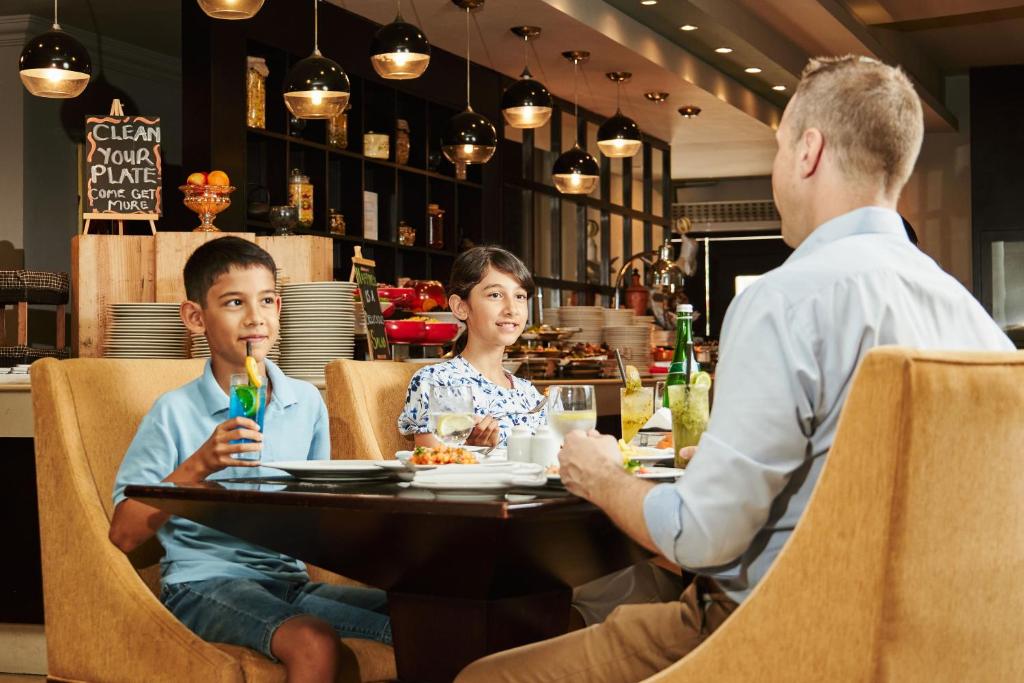 Bērni ēd maltīti restorānā, priecīgi sarunājoties ar pieaugušo, kam uz galda ir dažādi dzērieni un ēdieni. Restorāna interjers ir moderns un gaisīgs.