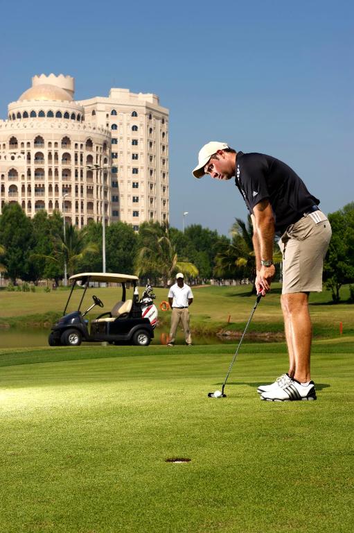 Golfists puttē uz zaļā laukuma ar modernu ēku fona, kamēr golfa mašīna un otra persona novēro procesu.