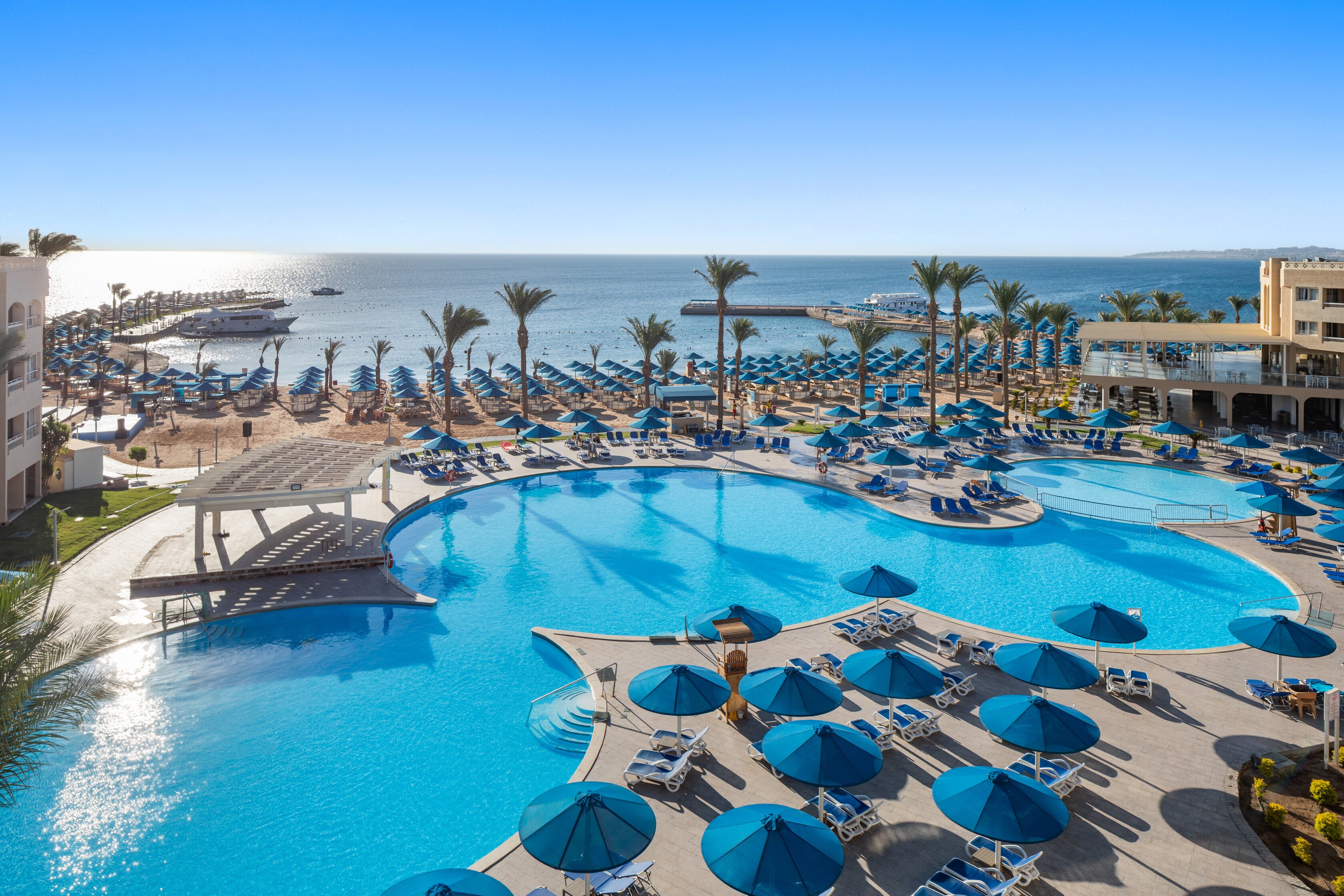 Pludmales skats no Beach Albatros Resort Hurghada ar baseinu un sauļošanās krēsliem.