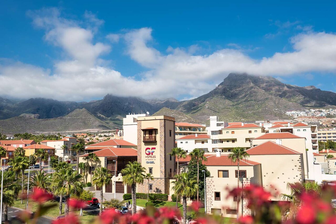 Skats uz viesnīcu "GF Hoteles Isabel" Tenerifē, ar kalniem fona, zilo debesi un palmu rindām priekšplānā. panoramic view with beach and pools