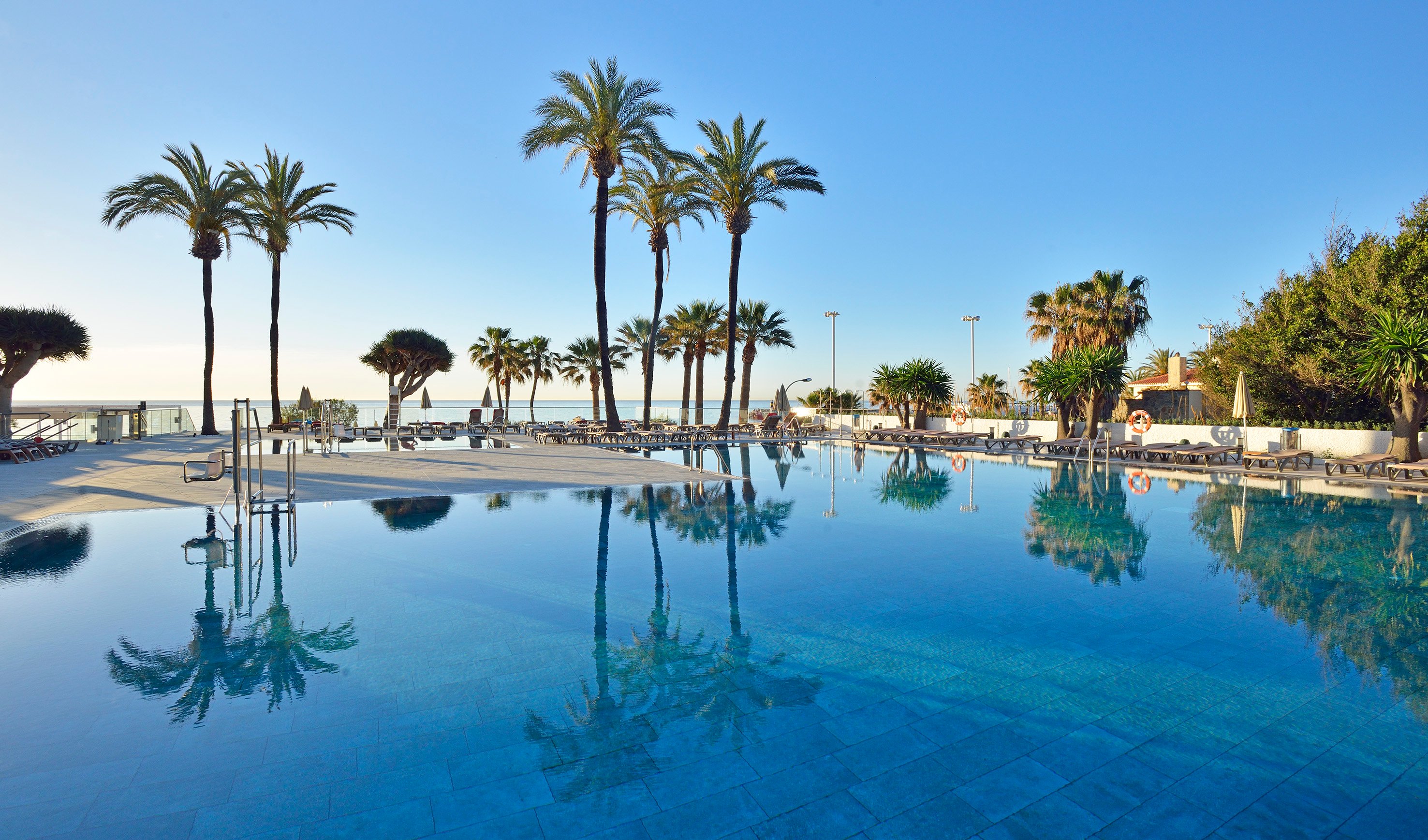 Lieliska pludmales baseina ainava ar palmām un saulainu debesu fonu, kas atspoguļojas ūdenī. Ideāla vieta atpūtai un relaksācijai. panoramic view with beach and pools