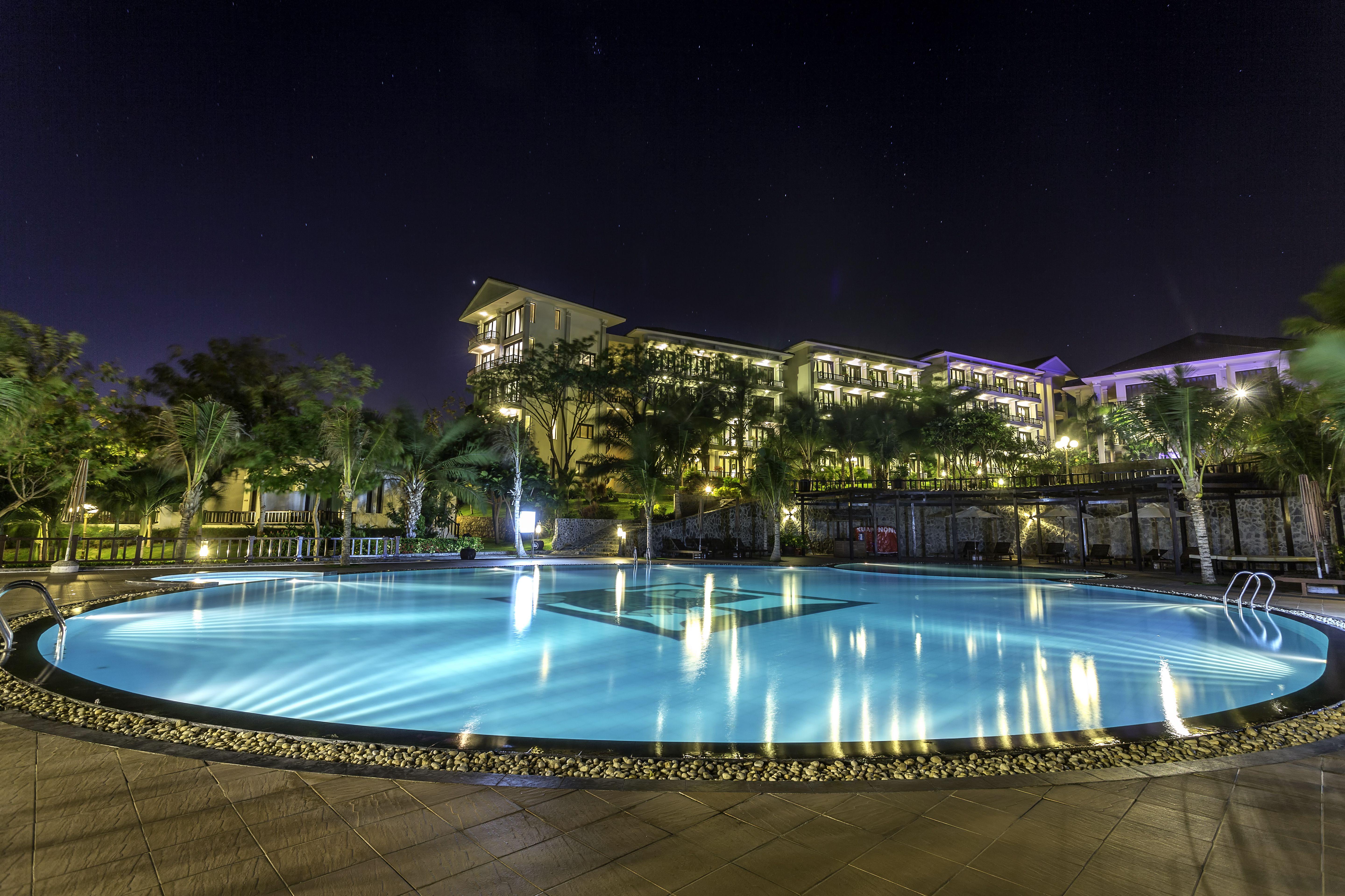 Peldbaseins ar apgaismojumu naktī, apkārt augstkokiem un viesnīcas ēkai. Klusa un relaksējoša atmosfēra. panoramic view with beach and pools