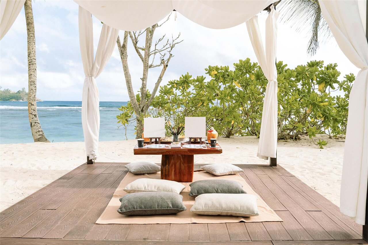 Kempinski Seychelles Resort privāta gleznošanas vieta pludmalē ar ērtiem spilveniem un skatu uz okeānu.