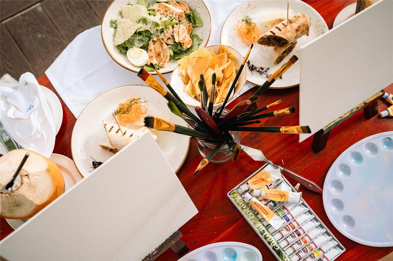 Gleznošanas piederumi, otas, krāsas un gardas uzkodas uz koka galda radošai atpūtai Kempinski Seychelles Resort.