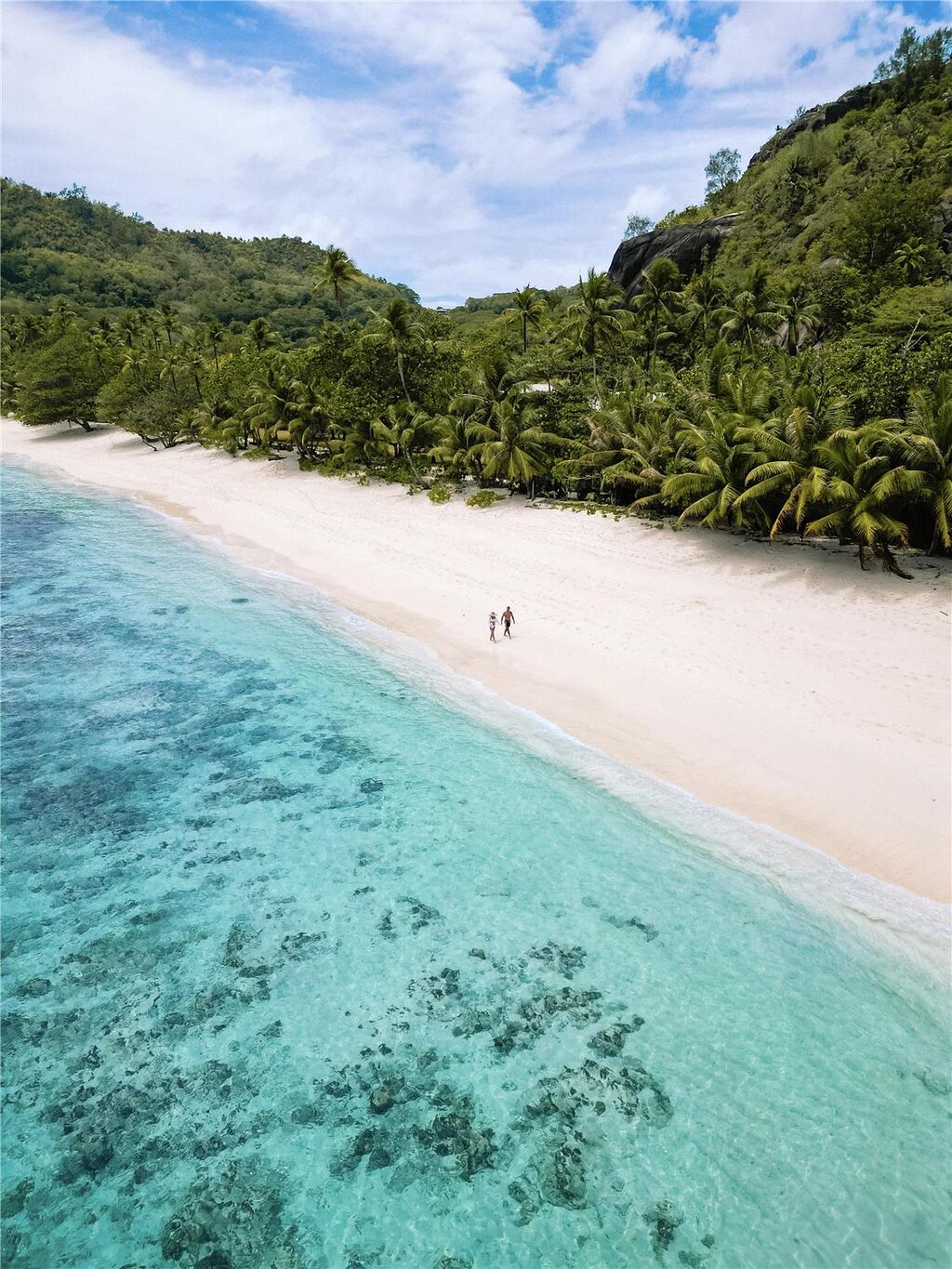 Pāris pastaigājas pa Kempinski Seychelles Resort balto smilšu pludmali pie tirkīzzila okeāna.