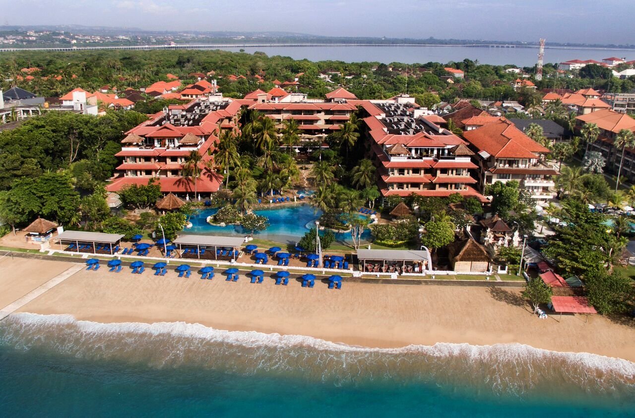 Tropu kūrortvieta ar baseinu, pludmali un zilu saulessargu rindu. Apkārt ir zaļa daba un sarkano jumtu ēkas. panoramic view with beach and pools