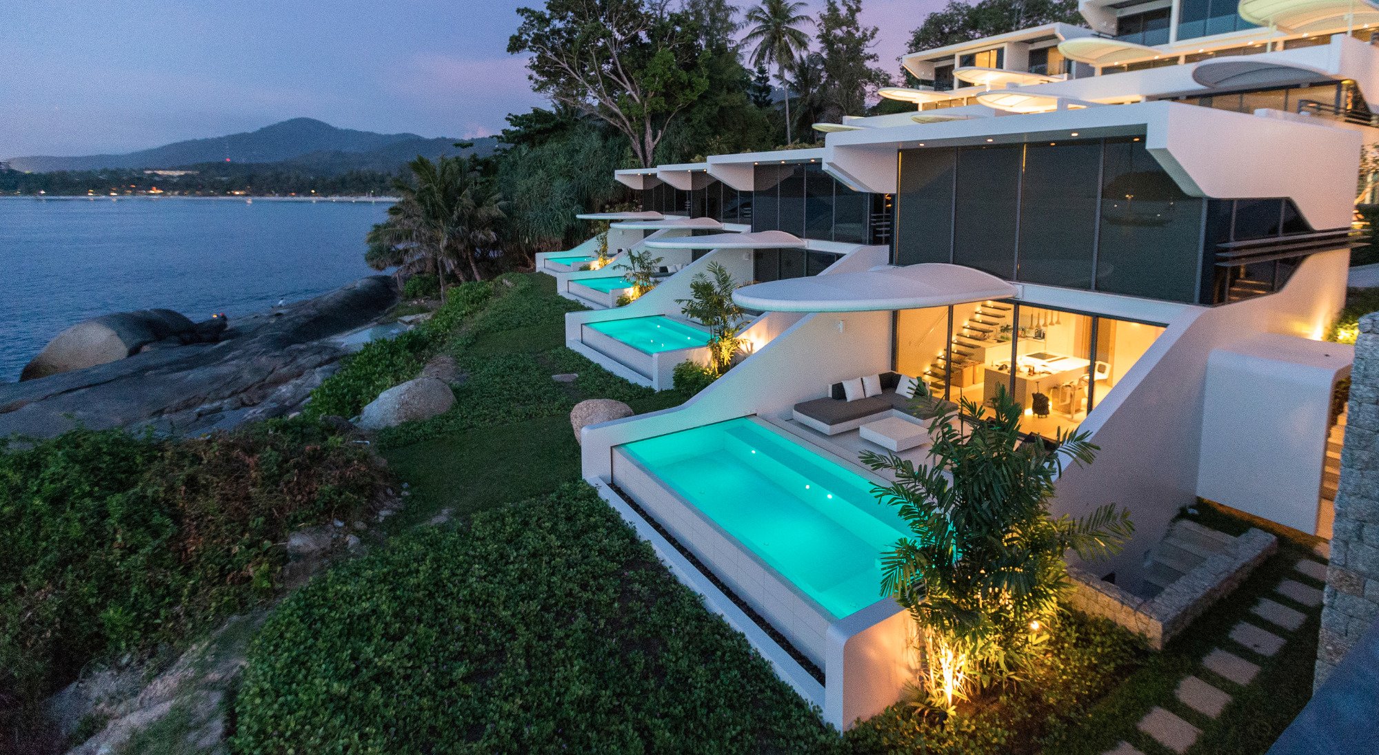 Mūsdienīgas villas ar privātiem baseiniem pie jūras, apgaismotas vakara gaismā, ieskautas krūmiem un palmām. panoramic view with beach and pools