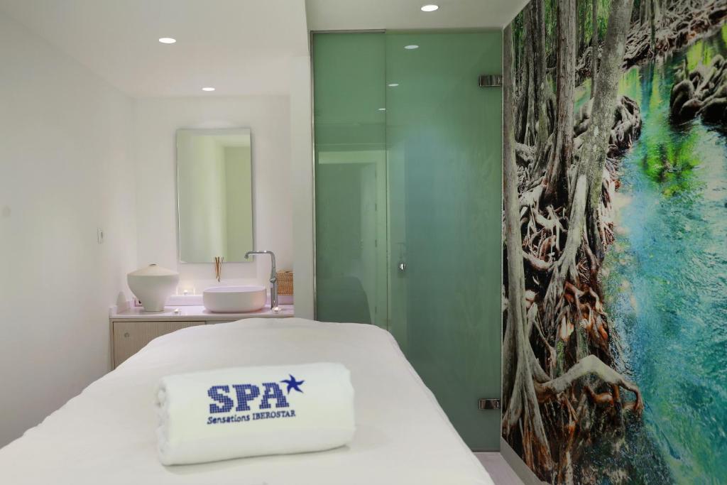 Spa telpa ar mūsdienīgu dizainu, balto masāžas galdu un krāšņu dabas sienu. Svaiga atmosfēra un relaksējoša vide, ideāli piemērota atpūtai un pašapkalpošanās brīžiem.