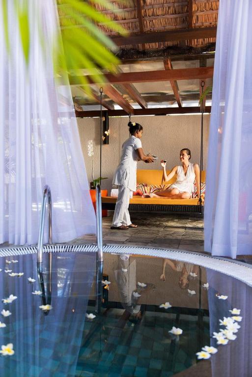 Relaksējošs spa brīdis, kur sieviete saņem dzērienu no masāžas terapeita, kamēr atpūšas pie baseina ar ziedlapiņām.