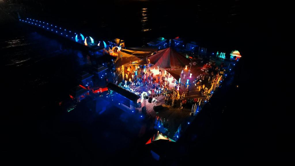 Krāsains un dzīvīgs festivāls pie ūdens, kur cilvēki pulcējas zem telts un izbauda gaismas šovu, kas atspoguļo jautru atmosfēru.