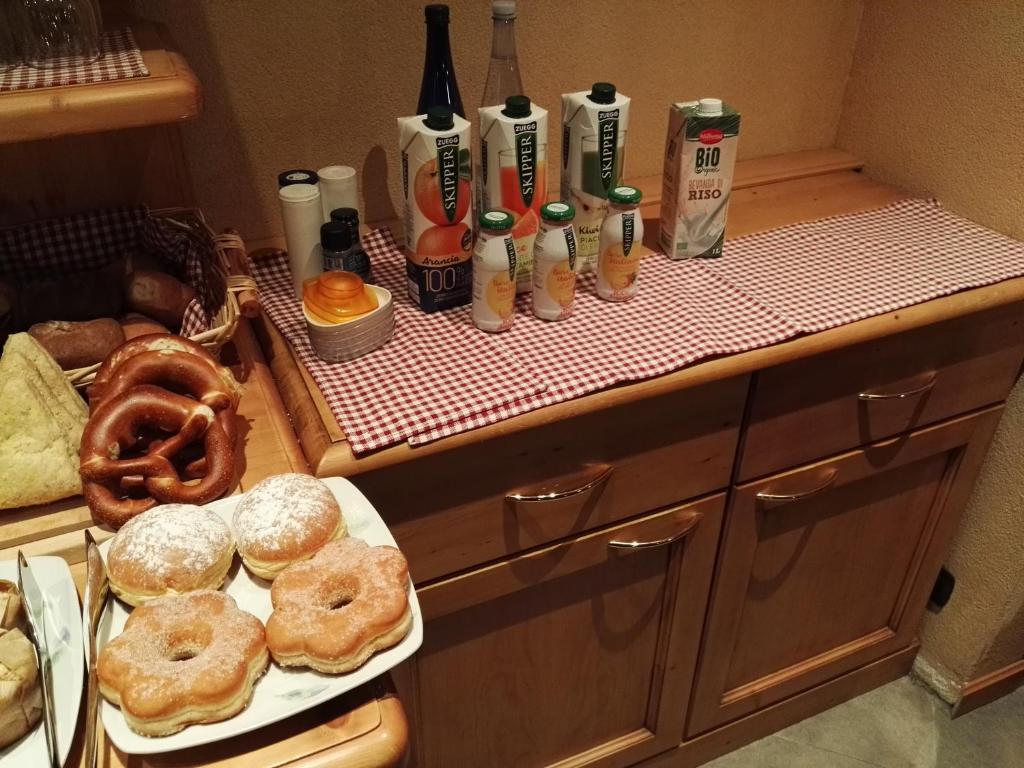 Broka ar dažādiem dzērieniem un saldumiem, tostarp donutām un maizes izstrādājumiem, uz koka galda ar sarkanu un baltu pārklāju.