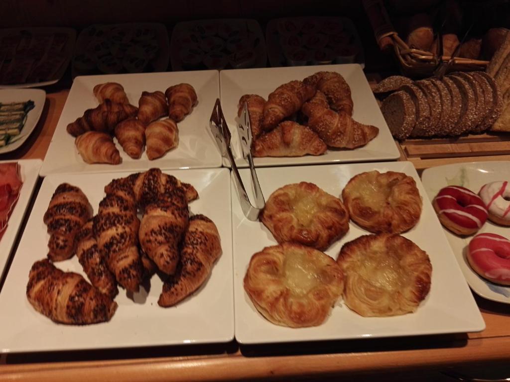Svaigi cepti kruasāni un saldētas maizītes, pasniegtas uz baltām šķīvēm. Uz galda redzami dažādi maizes izstrādājumi un saldumi.
