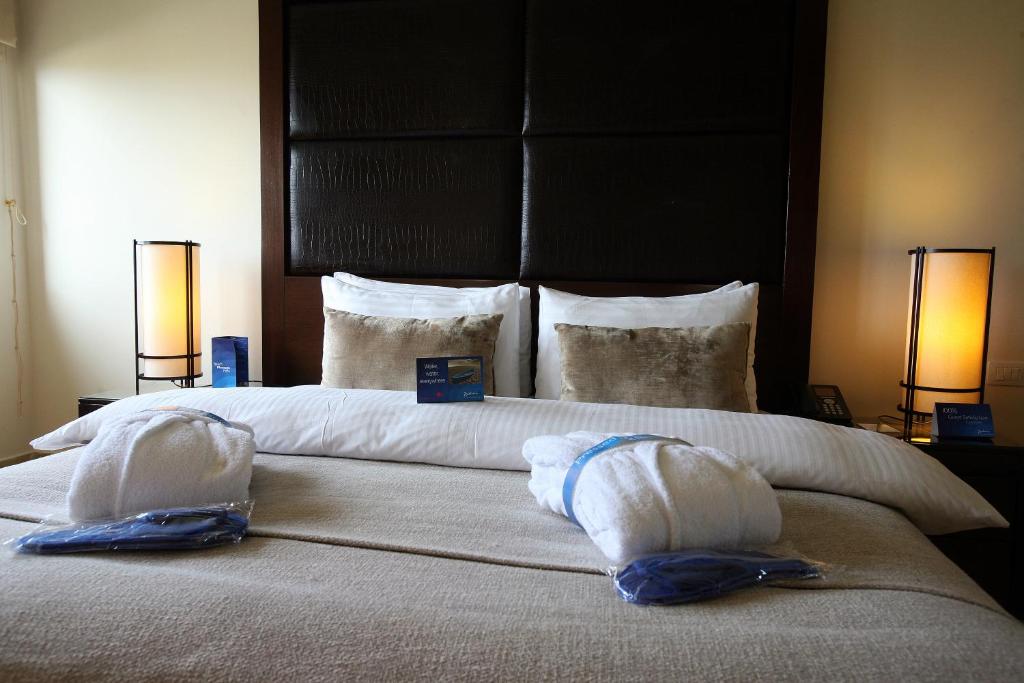 Elegants viesnīcas numurs ar divguļamo gultu, mīkstām spilveniem un rūpīgi salocītām dvielim. Gultas galvas daļā ir tumša paneļa apdare, un blakus atrodas naktslampas.