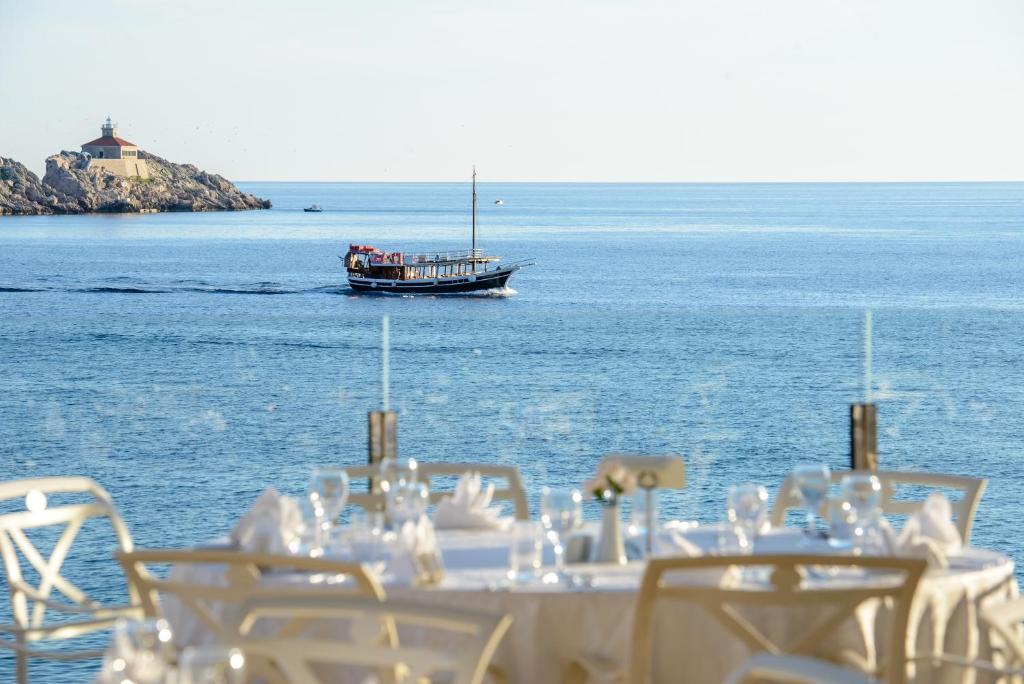 Skats uz jūru ar tradicionālu laivu, kas peld gar krastu, un restorāna galdiem priekšplānā.