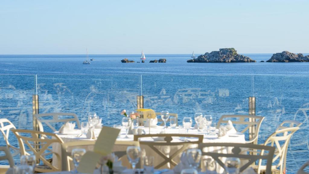 Skats uz mierīgu jūru no restorāna ar elegantām galdām un krēsliem, kas izvietoti uz terases. Tālā redzami kuģi un akmeņi ūdenī, radot relaksējošu atmosfēru.