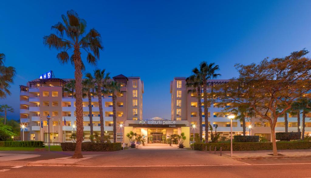 Moderns viesnīcas ēka ar palmām un nakts apgaismojumu, kas rada mierīgu atmosfēru. Viesnīcas nosaukums redzams virs ieejas.