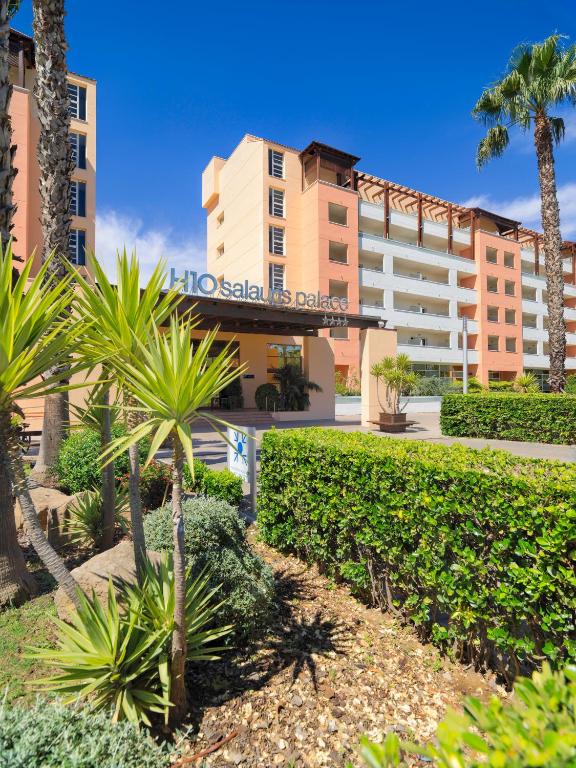H10 Salou Palace viesnīcas ieeja ar mūsdienīgu arhitektūru un zaļu ainavu, kas veidota ar palmām un krūmiem. Ideāla vieta atpūtai un atvaļinājumam.