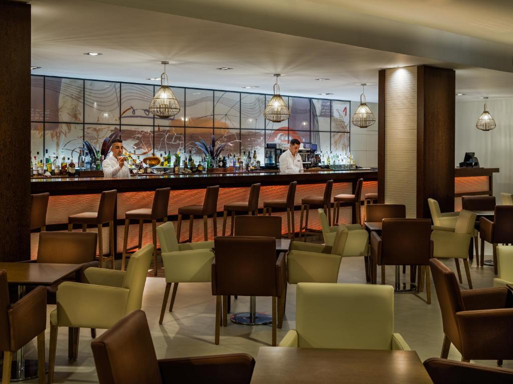 Mūsdienīgs bārs ar elegantiem krēsliem un izsmalcinātu apgaismojumu, kur divi bārmeņi gatavo dzērienus. Fons ar stilizētu mākslu un plašu dzērienu izvēli.