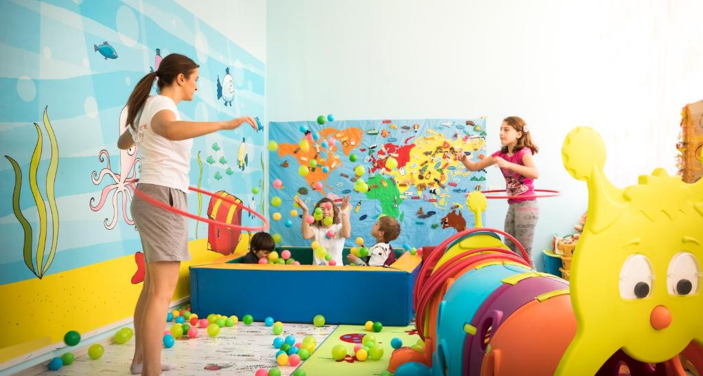 Bērnu rotaļu telpa ar spilgtiem sienu zīmējumiem, kur bērni spēlē ar krāsainām bumbiņām un rotaļlietām, kamēr pieaugušais virsū griež hula gredzenu.