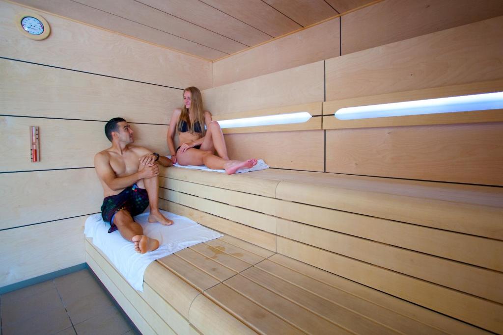 Pāris relaksējas modernā saunā, sēžot uz koka soliņiem. Telpa ir gaiša un mūsdienīga, ar temperatūras un mitruma rādītājiem uz sienas.