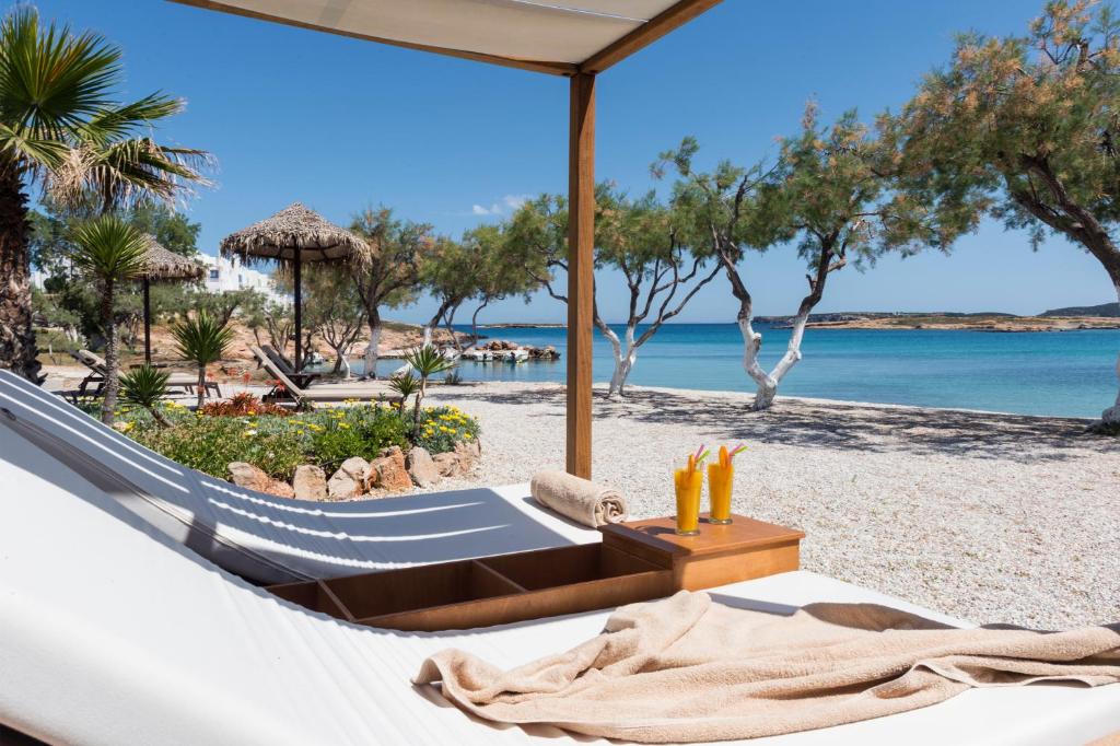 Saules gaisma izgaismo pludmali ar palmām un koka lapenēm, kur atrodas atpūtas krēsli un kokteiļi. Aiz krēsliem redzams dzidrs ūdens un smiltis, radot relaksējošu atmosfēru.