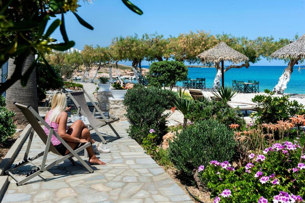 Atpūtas zona pie pludmales ar sauļošanās krēsliem, ziedošiem augiem un palmām, kur sieviete atpūšas ar grāmatu rokās. Jūras skats un saulaina debesis papildina mierīgu atmosfēru.