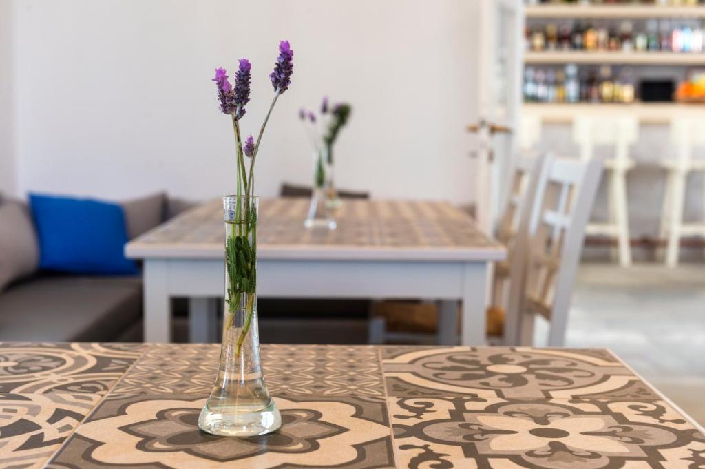 Violeta ziedu pušķis stikla vāzē uz galda ar rakstainu virsmu, blakus gaišai krēslai un kafejnīcas interjeram.