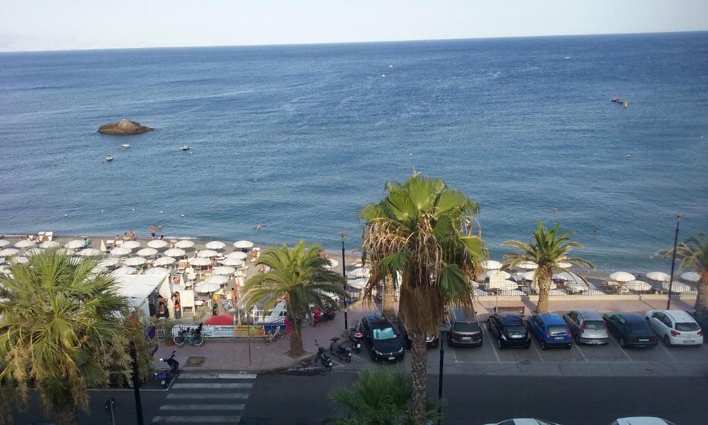 Krāšņs pludmales skats ar palmām, saulessargiem un cilvēkiem, kas atpūšas pie ūdens. Jūra ir mierīga, un krastā redzamas automašīnas.