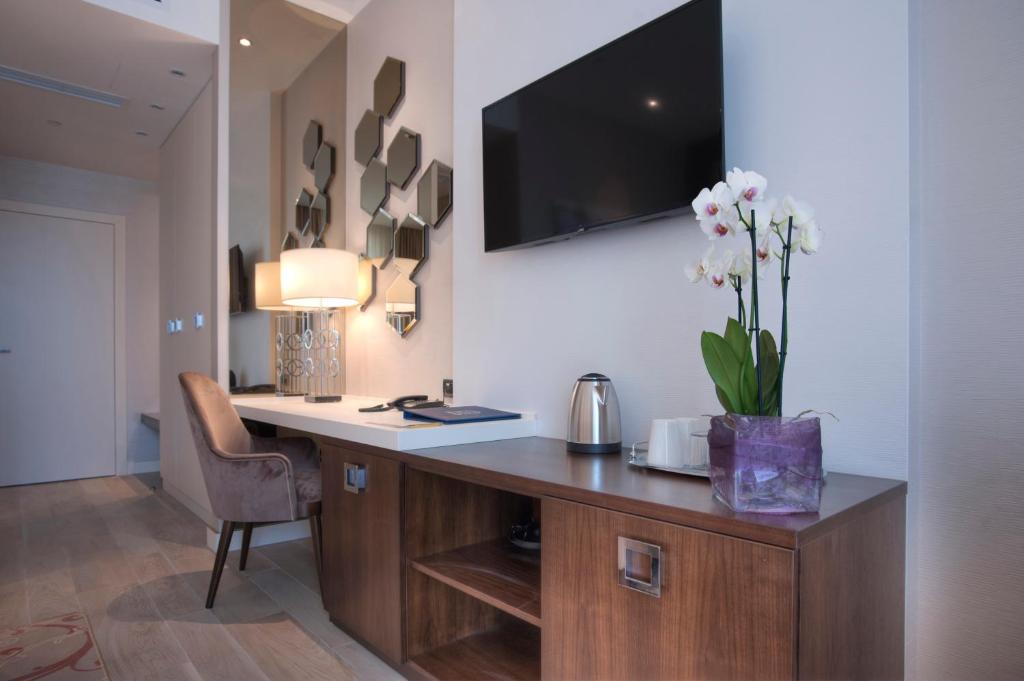 Mūsdienīgs viesnīcas numurs ar elegantu galdu, krēslu, televizoru un dekoratīvām spoguļu plāksnēm uz sienas. Uz galda atrodas puķu pods ar orhidejām un tējas komplekts.