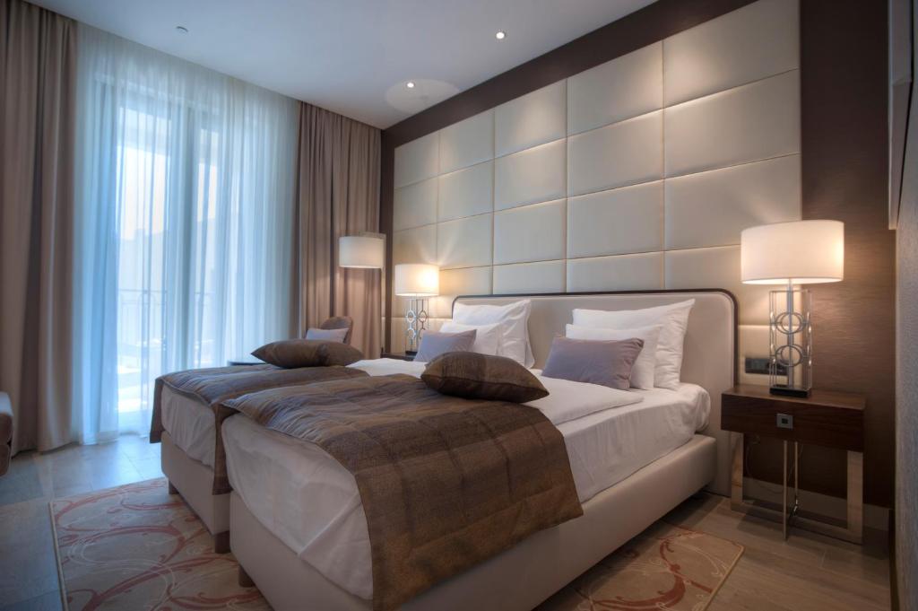 Mūsdienīga viesnīcas guļamistaba ar divām gultām, mīksto apgaismojumu un lieliem logiem, kas nodrošina dabiska apgaismojuma plūsmu. Elegants interjers ar neitrālām krāsām un teksturētām sienām.