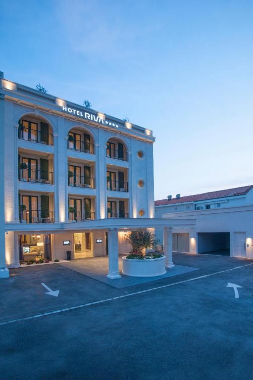 Elegants viesnīcas ēkas eksterjers ar modernu arhitektūru un apgaismojumu, kas radīts vakara atmosfērā. Viesnīca Riva piedāvā komfortu un stilīgu dizainu.