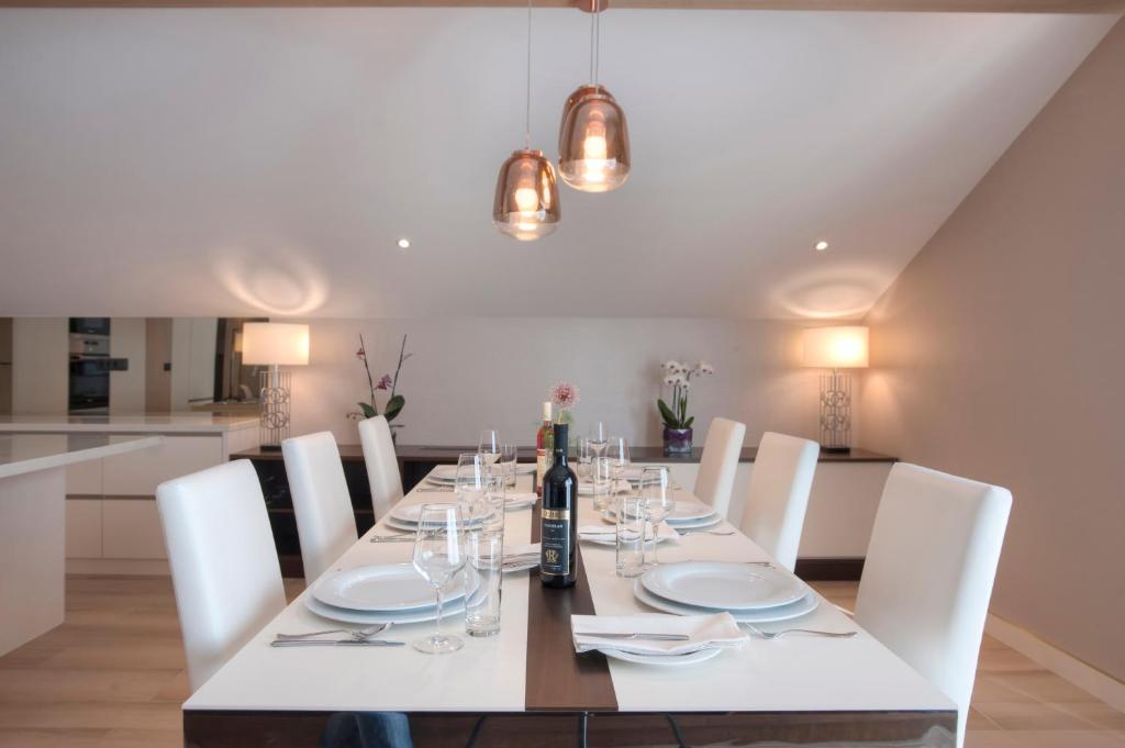 Moderns un elegants ēdamistabas interjers ar garu galdu, kas sagatavots viesiem. Galda centrā ir pudele vīna un glāzes, apgaismojums veido siltu atmosfēru.